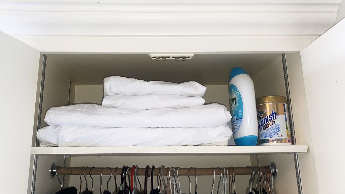 Top of wardrobe laundry shelf showing white bed linen folded next to laundry detergent.