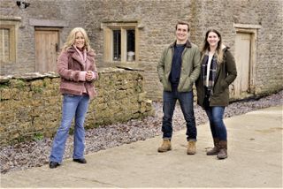 Sarah Beeny stands outside a stone cottage in the country, with a low stone wall and gravel on the ground, with young couple James and Jodie