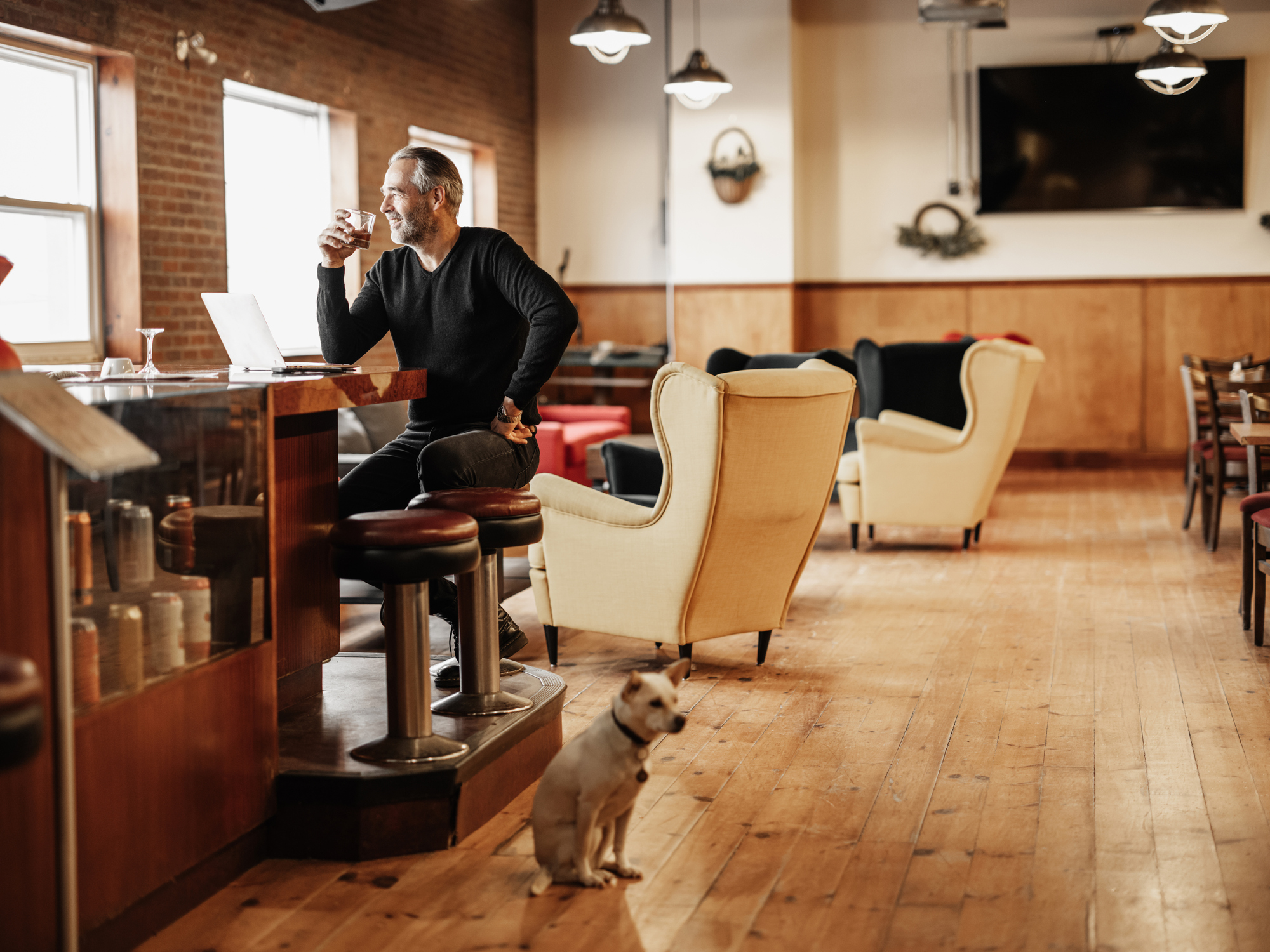 Owner of small business working at the bar, working on lap top while having a drink. Dressed in casual clothes with black sweater and jeans. Interior of vintage bowling alley in the morning with natural light. Small dog in the foreground.