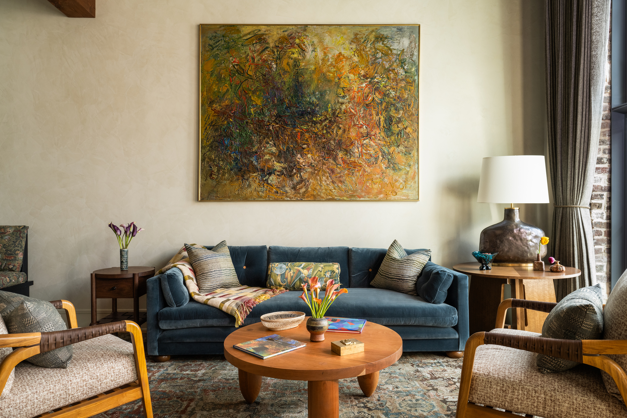 modern living room with blue velvet sofa, large artwork, plaster walls, round timber coffee table, two gray and timber armchairs, a patterned rug, and a coffee table and lamp