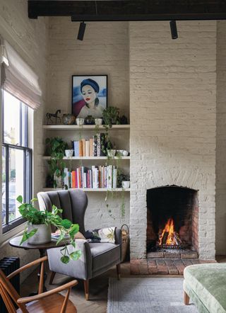 White brick fireplace with a fire lit inside of it. There is a gray accent chair with a round side table next to it. On the side table is a green plant and a shelf of books is on the wall behind it.