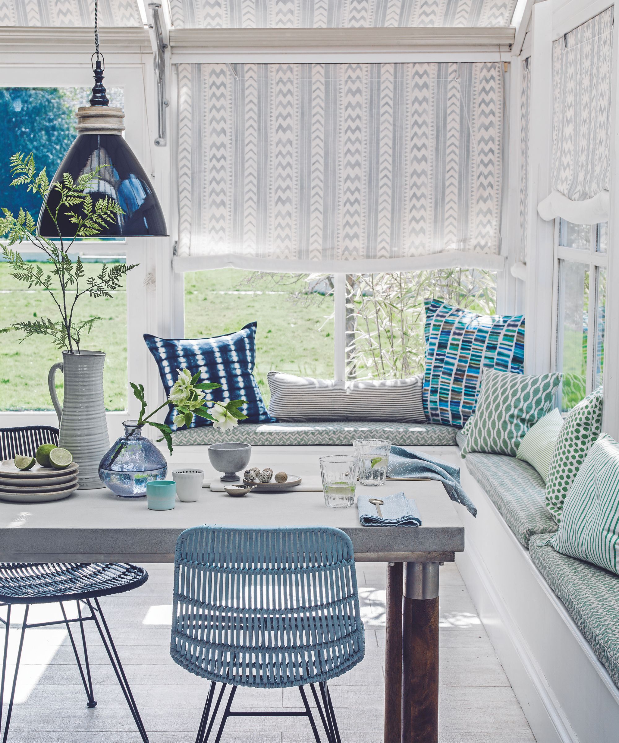 A bright and sunny blue and white sunroom with green banquette seating and colorful, mismatched cusions. There is a large table laid with blue napkins, plates and vases, with a large white jug with a green leafy stem inside. Behind are large windows with white and grey patterned blinds half closed.