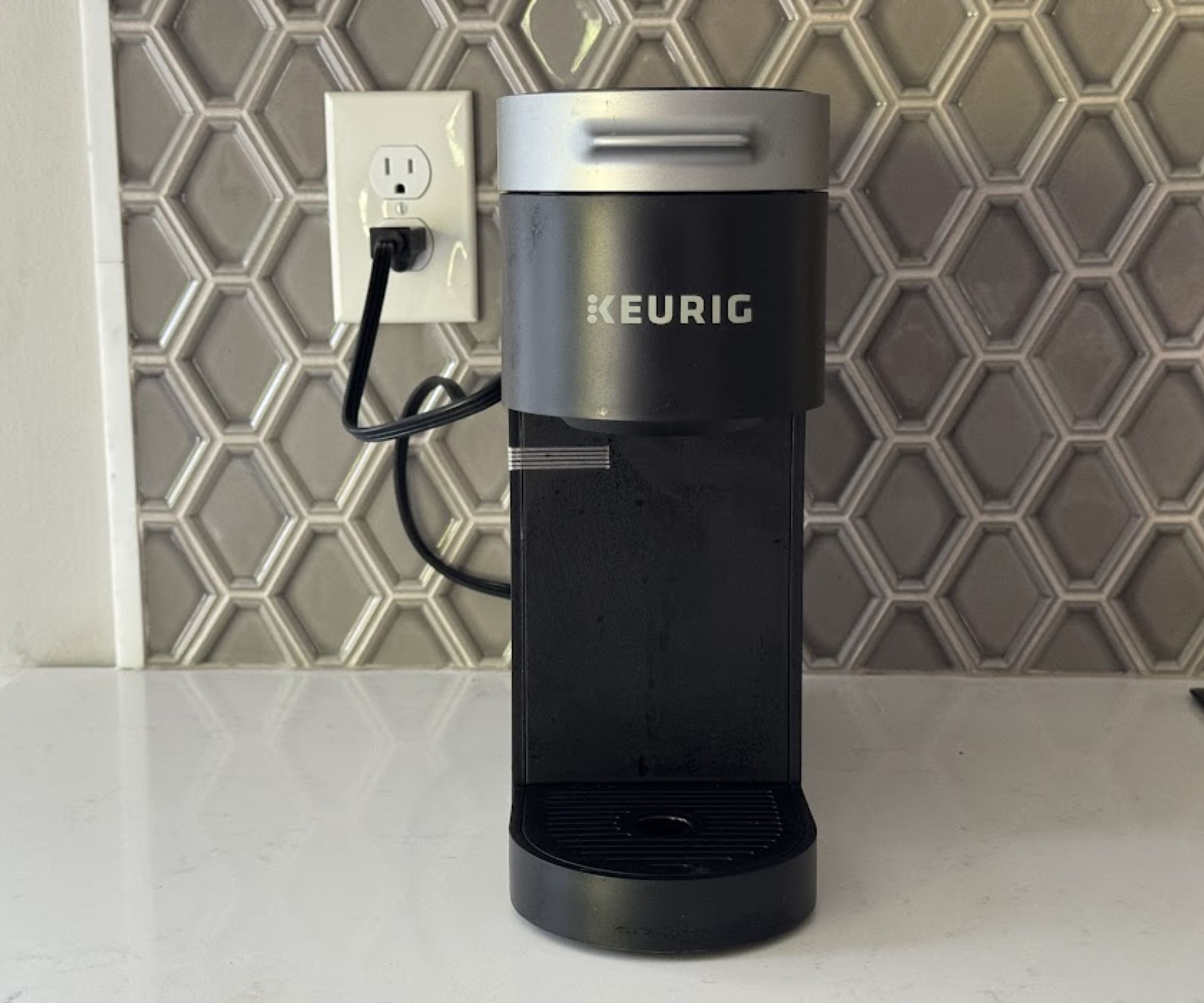 A black Keurig coffee machine plugged into a wall outlet, in front of a grey tile backsplash