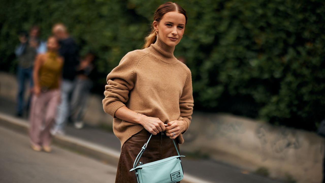 Guest at milan spring 2025 fashion week wearing beige turtleneck sweater and blue handbag 