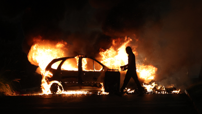 A protester walks by a burning car at night 