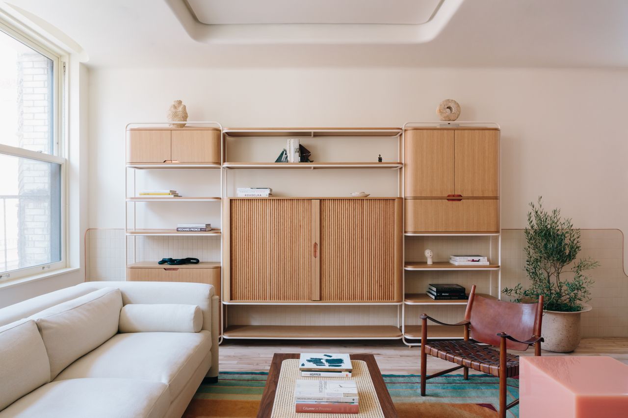 A lounge area with a white sofa, a rectangular wooden coffee table, a leather chair and a large console with many levels for storage.