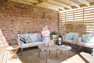 woman sitting in outdoor pergola area on gray table set, with wooden slat wall and a brick wall behind her