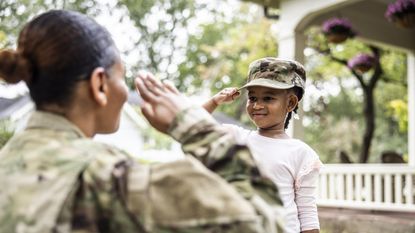 A soldier salutes her young daughter, who's wearing the soldier's uniform cap.
