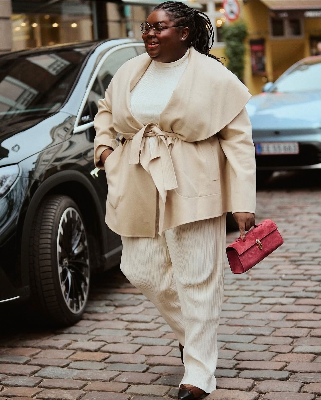 Abi wears an ivory coat, ivory pants, black flats, and a red Savette bag.