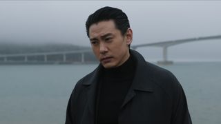 Jang stands by the water's edge with a bridge behind him in The Recruit season 2