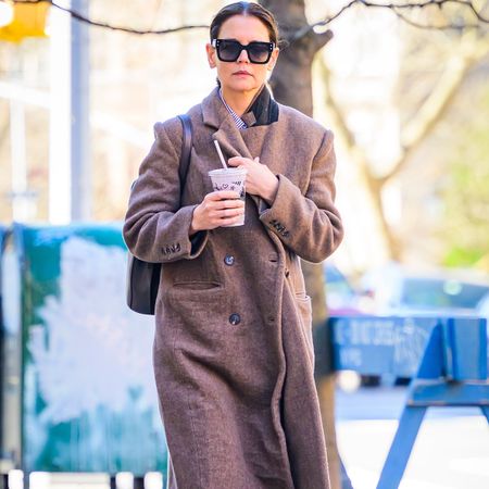 Katie Holmes wears a long brown coat, gray sweatpants, and gray sneakers.