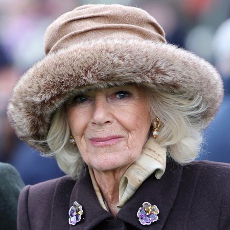 Queen Camilla wearing a fur hat and purple coat with flower brooches in 2025