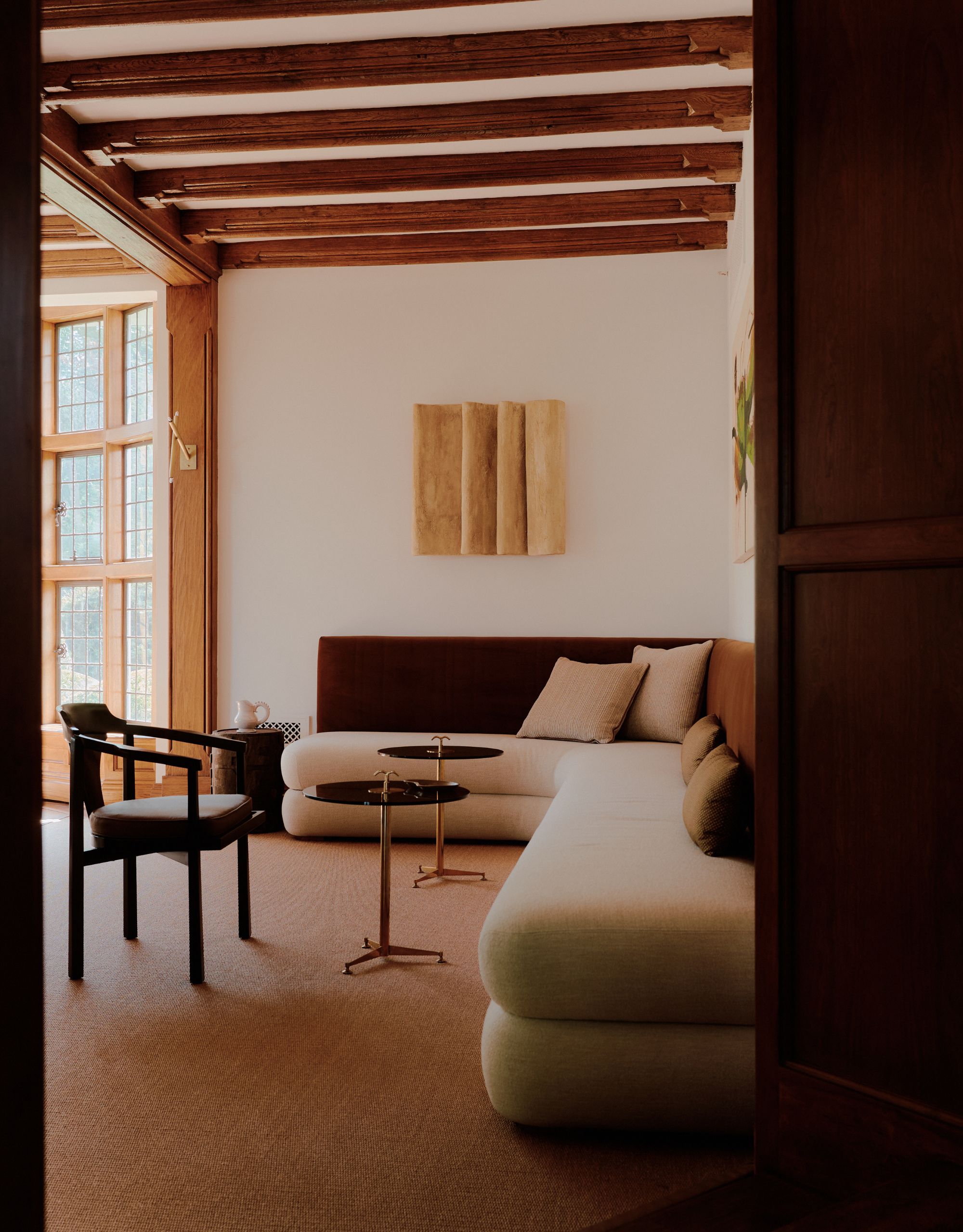 A banquette seating area with a rust brown velvet back and cream linen seating with beige and brown cushions in a neutral space with wooden beams on the ceiling and artwork on the walls