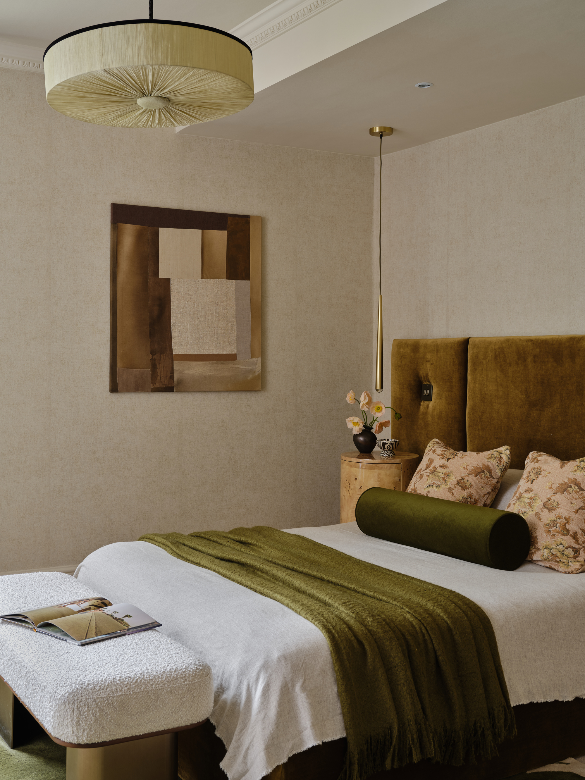 Image of a neutral bedroom with a ochre velvet headboard and olive green pillows and throws on the bed. There is a brown painting on the wall and a boucle bench seat at the end of the bed.