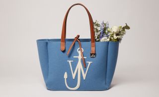 View of a blue JW Anderson tote bag featuring brown buckle handles and the white 'JW' anchor logo. The bag has flowers and foliage inside and is pictured against a grey background