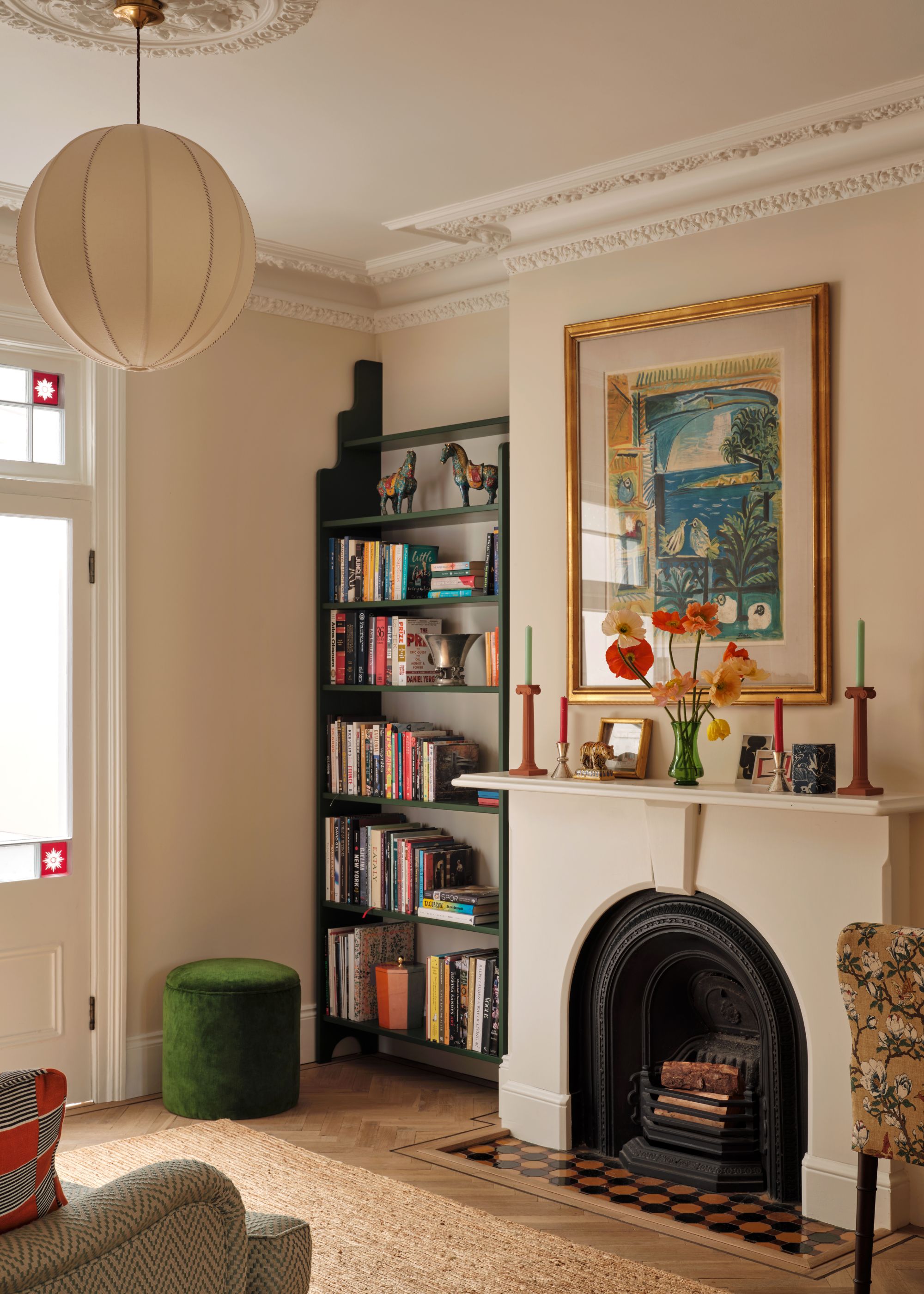 A corner of a living room with warm white walls, a white ceiling with cornicing details, a white fireplace with an abstract artwork above it, an alcove bookshelf painted dark green, and wood floors with a jute rug.