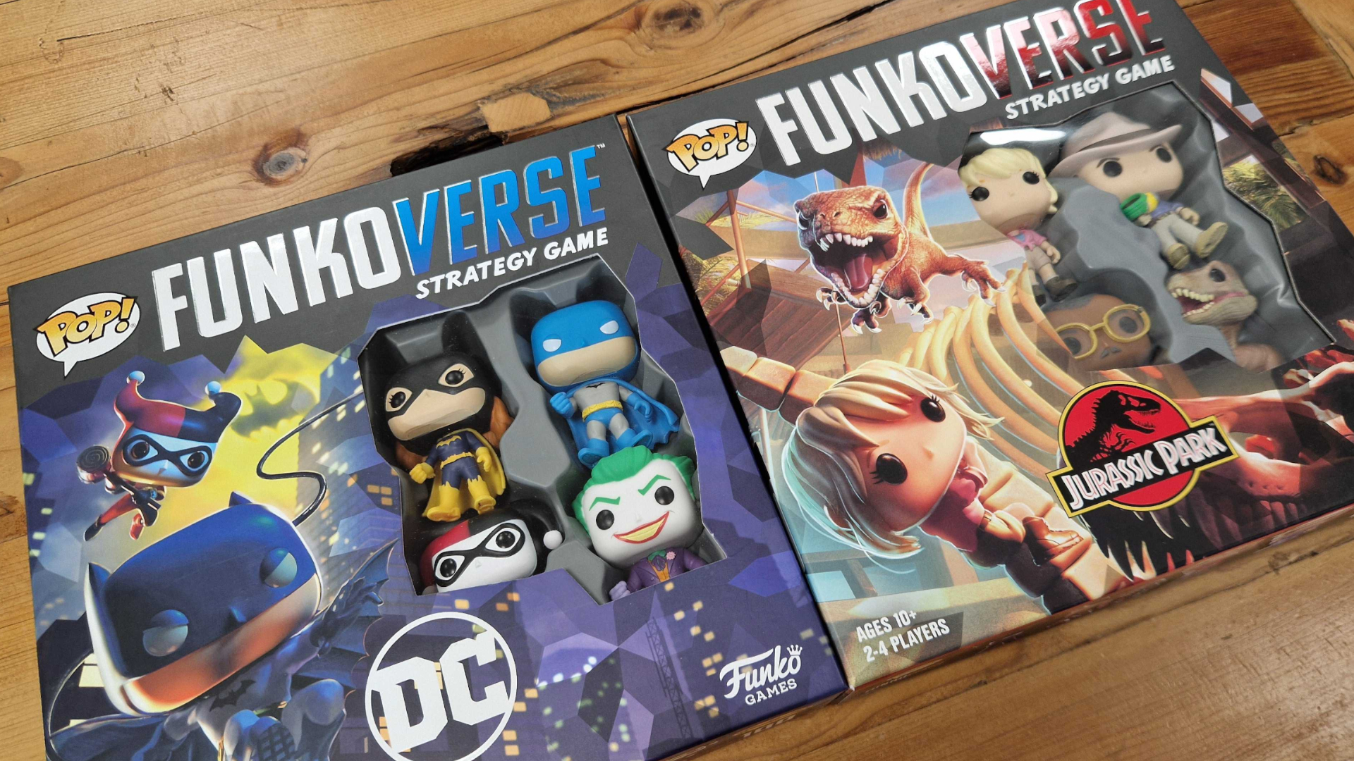 Funkoverse Strategy Game boxes for Batman and Jurassic Park laid out side by side on a wooden surface