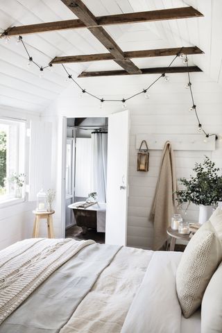 A white bedroom with shiplapped walls, coat hooks, overhead wooden beams and en suite bath in shot