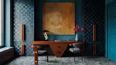 A dark blue drenched room with a wooden desk in the middle with drawers in between two chairs in the shape of an upside-down triangle with two fitted wardrobes either side of the desk and a large gold colored artwork hangs centrally above the desk which has a lamp and a vase of flowers on it as well as two books