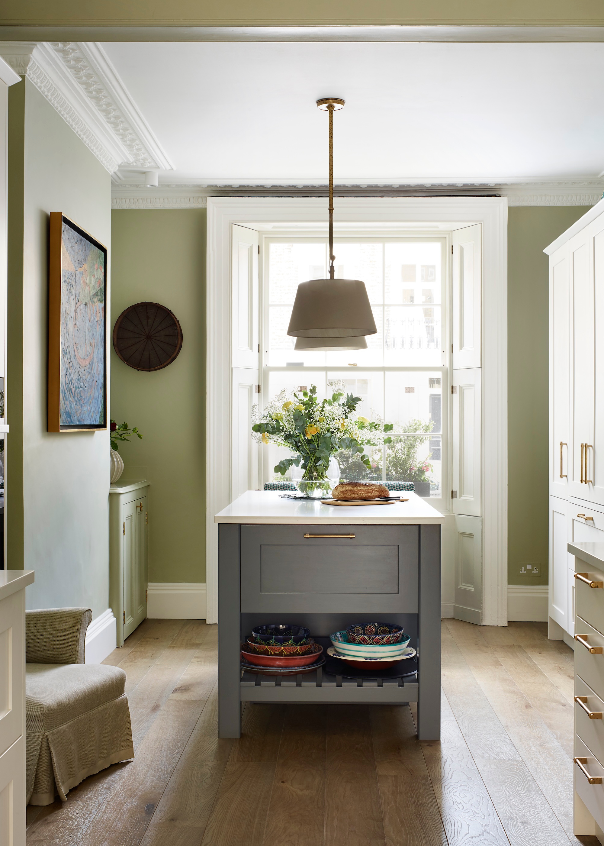 A neutral kitchen with earthy green-painted walls, a pendant light hangs above the kitchen island next to tall off-white cabinetry and there is a piece of art on the wall next to an armchair