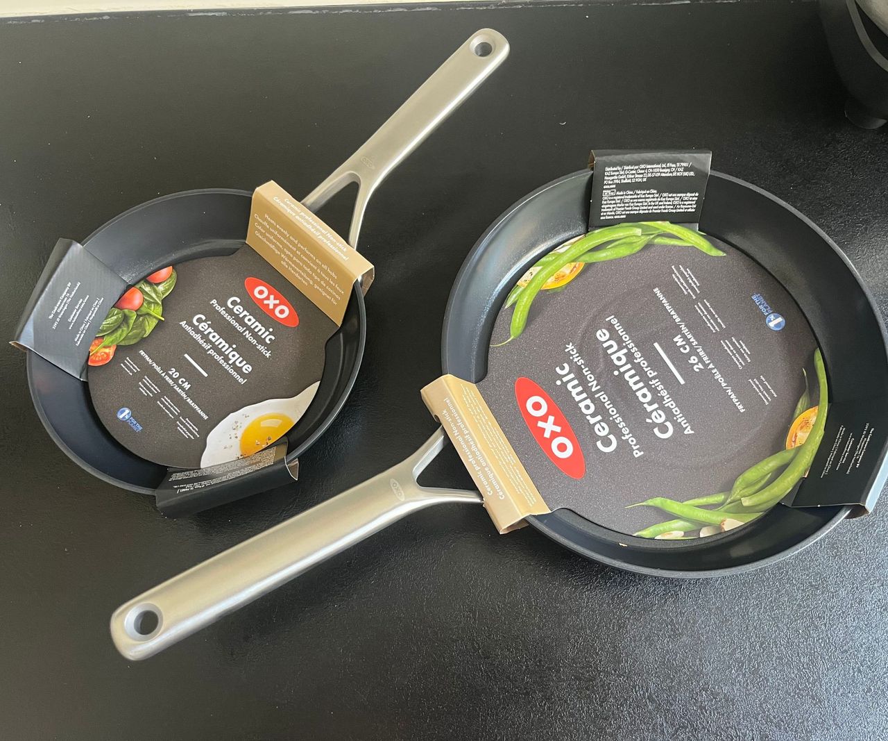 Two black ceramic frying pans on a black countertop with their OXO packadging sleeves on them.