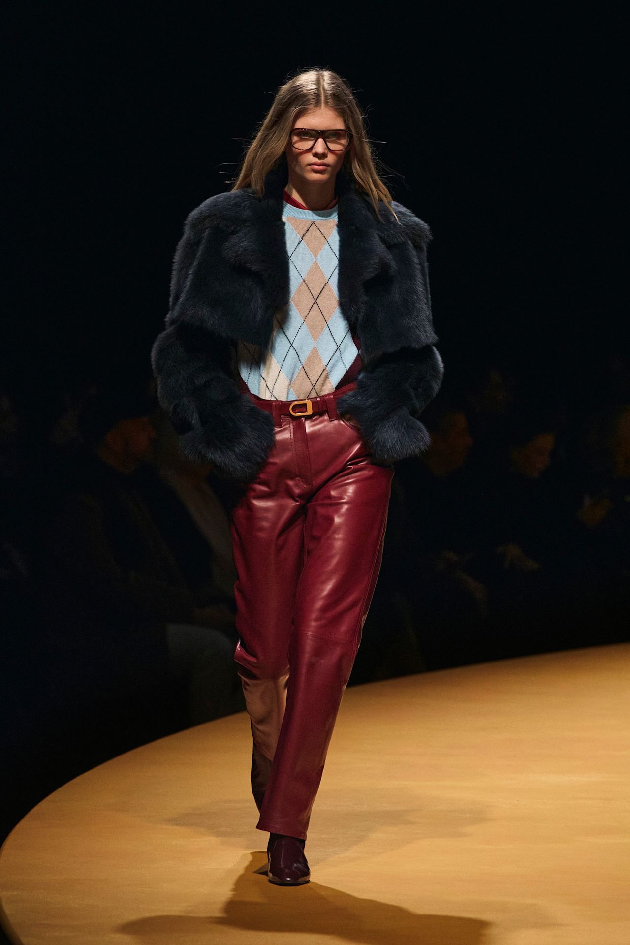 a model at Khaite Fall 2025 wearing a blue argyle sweater, black fur coat, and red leather pants