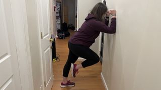 Trainer Jennifer Rizzuto performs a marching wall plank in a hallway. Her forearms rest against the wall; one of her feet is on the floor while the other is lifted and bent at the knee.