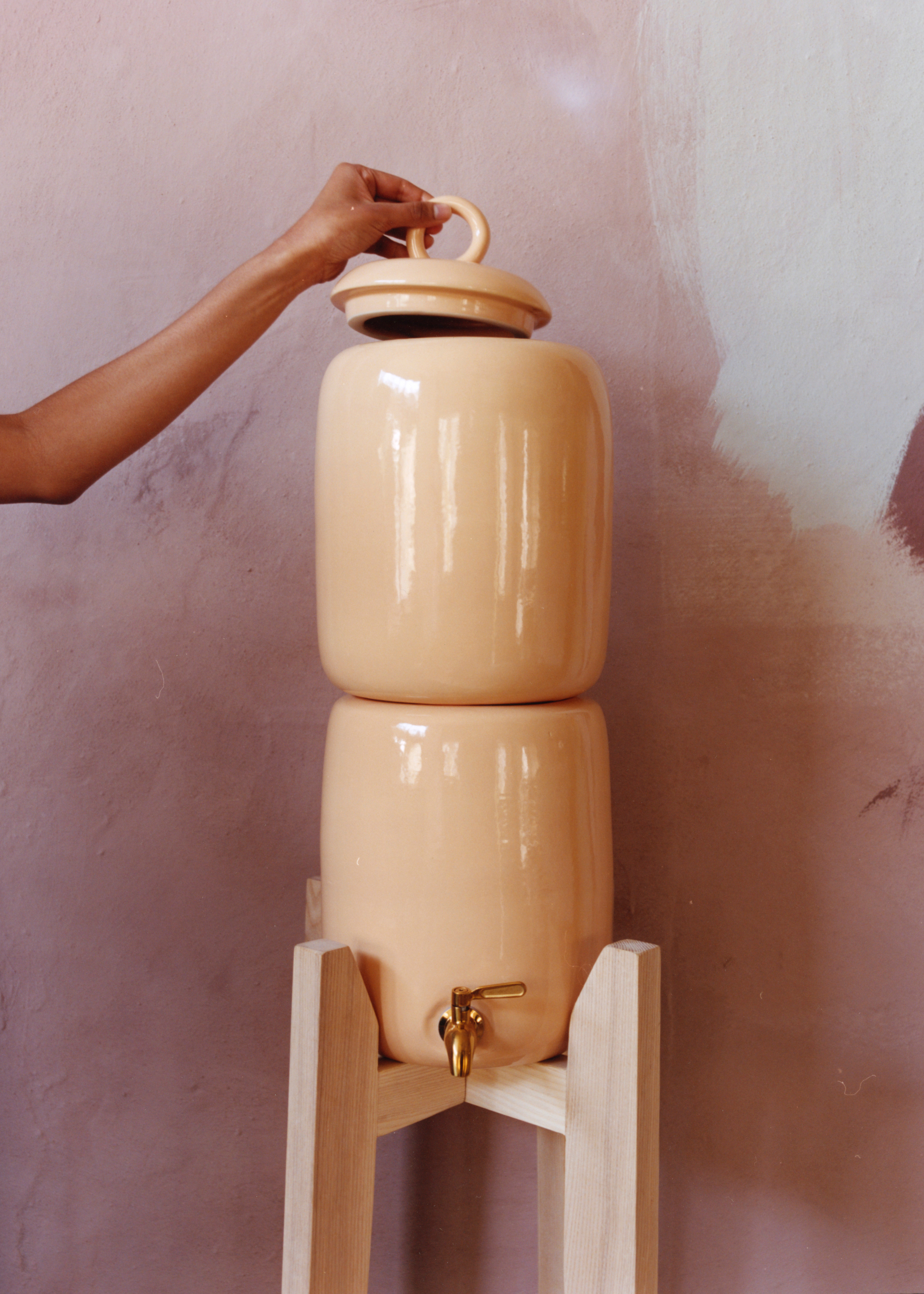 A beige ceramic glazed water vessel on a wooden stand with a hand opening the top