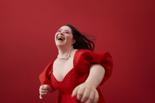 A young girl laughing while wearing a puffy sleeve red dress in front of a red background.