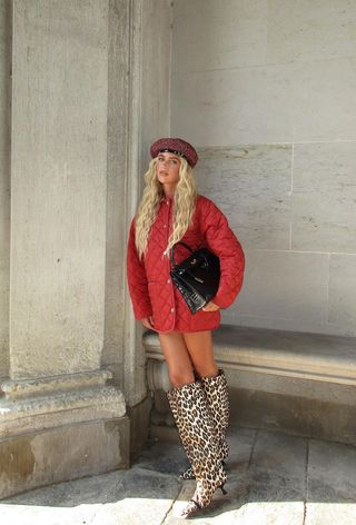 animal print boot trend is shown on a woman standing on the sidewalk wearing a red quilted jacket with leopard print knee-high boots, a black bag, and a red beret