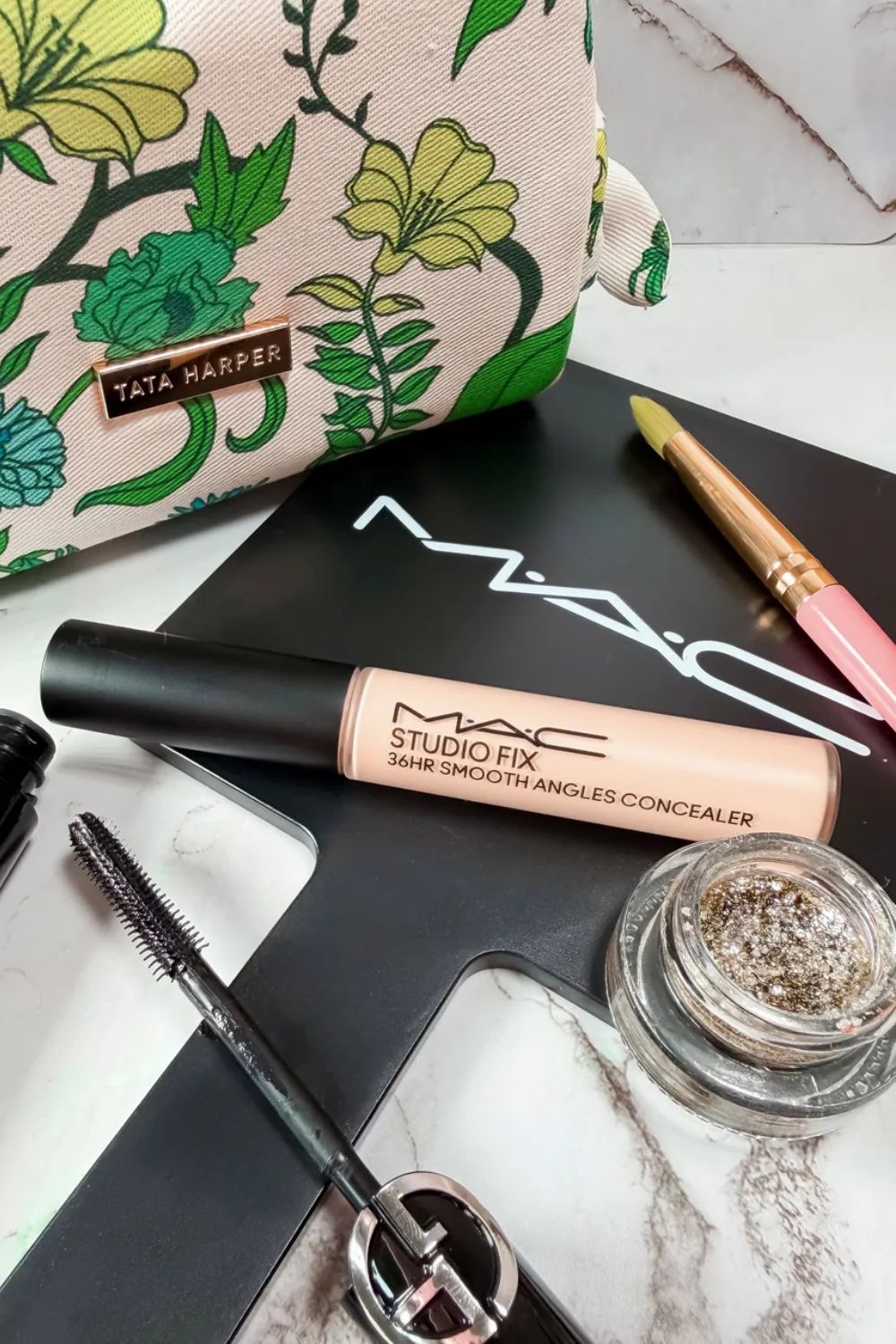 A flatlay of makeup products containing the MAC Studio Fix 36HR Smooth Angles Concealer, and a Gucci mascara and Tata Harper makeup bag
