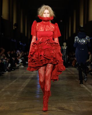 A model wearing a poppy red ruffled lace minidress with a ruff walking at the F/W 25 Alexander McQueen show.