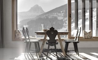 Close up interior view of a light wood dining table, dark wood chairs and black and white rug. Through the window there is a view of the snow covered landscape