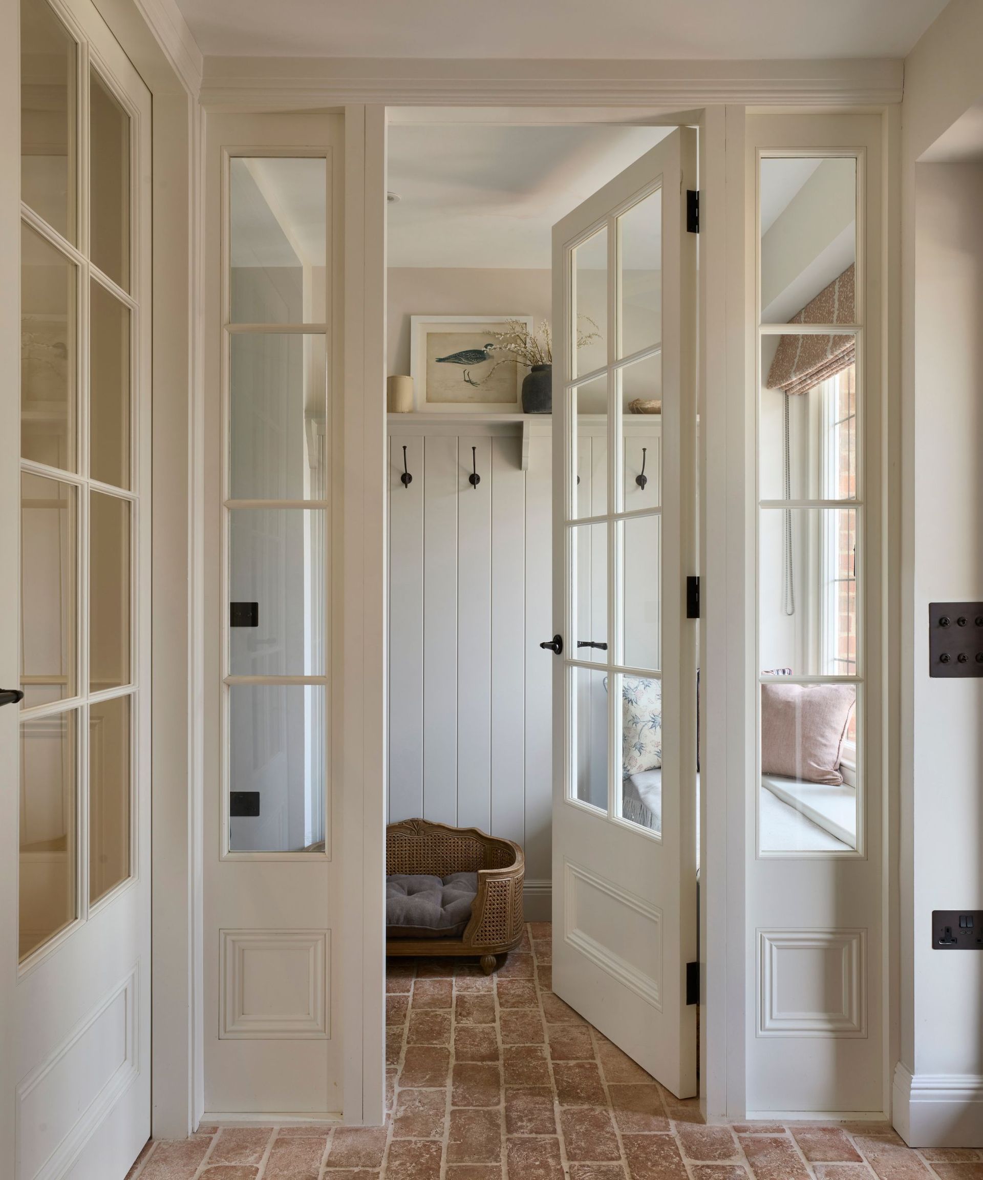 A white entryway with white doors opening up to a room with hooks and a pet bed, and brick tiles on the floor
