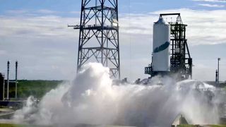 Outside on a sunny day, with some flat, white cloud cover, A small cylindrical tank with a large black feather painted on the side billows water and steam in a rush below. It is supported by a short metal structure, adjacent to a tall lighting tower.