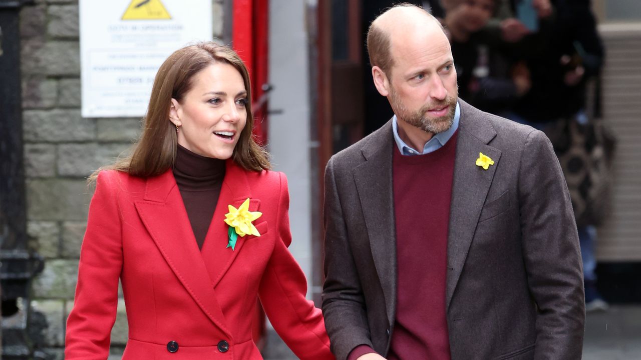 Kate Middleton wearing a red coat and yellow daffodil looking to the right walking outside next to Prince William, also looking at the same thing and wearing a daffodil on his blazer