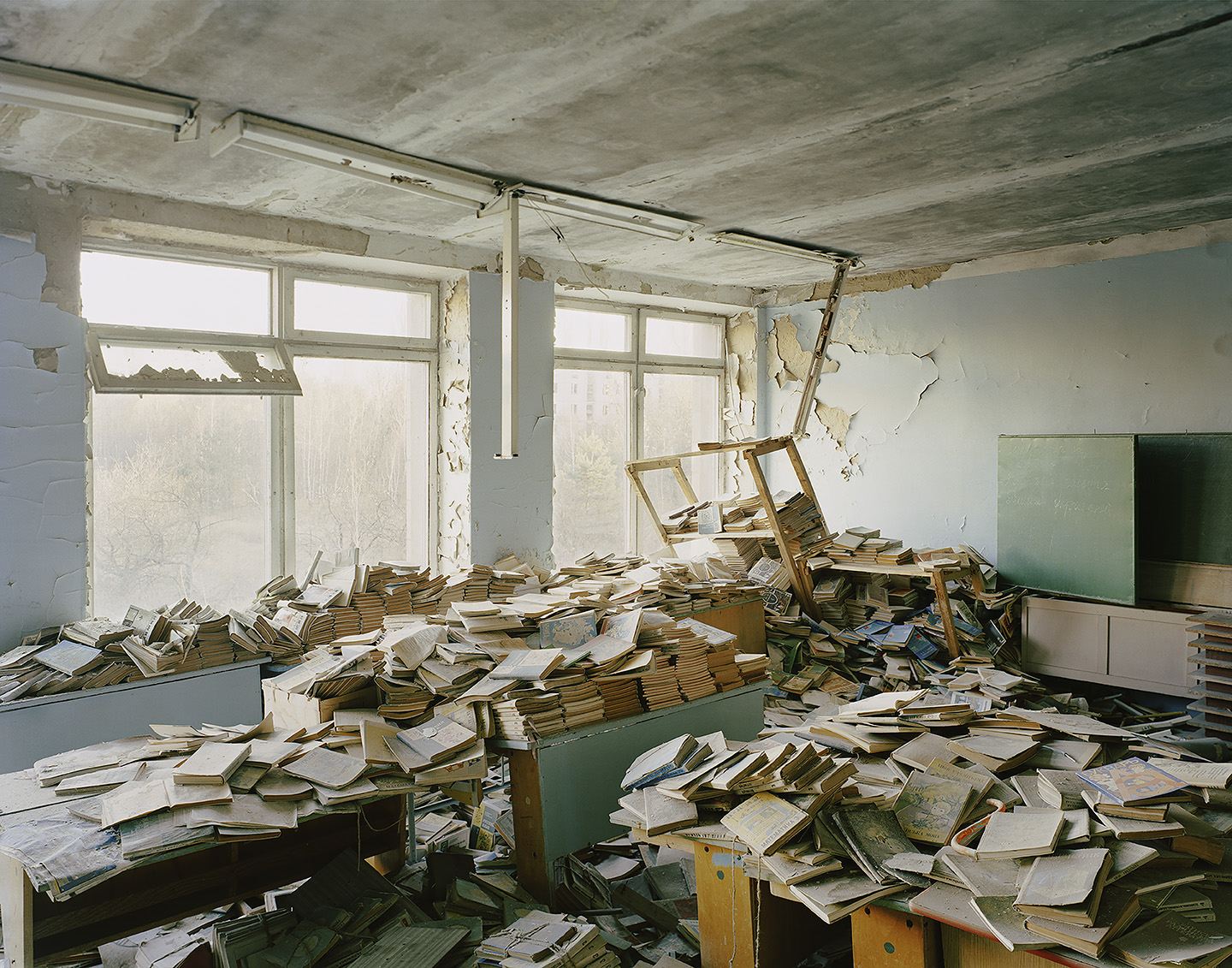 An abandoned school library in Pripyat, with mounds of dusty books piled across desks and floor beneath peeling walls and a sagging ceiling light.