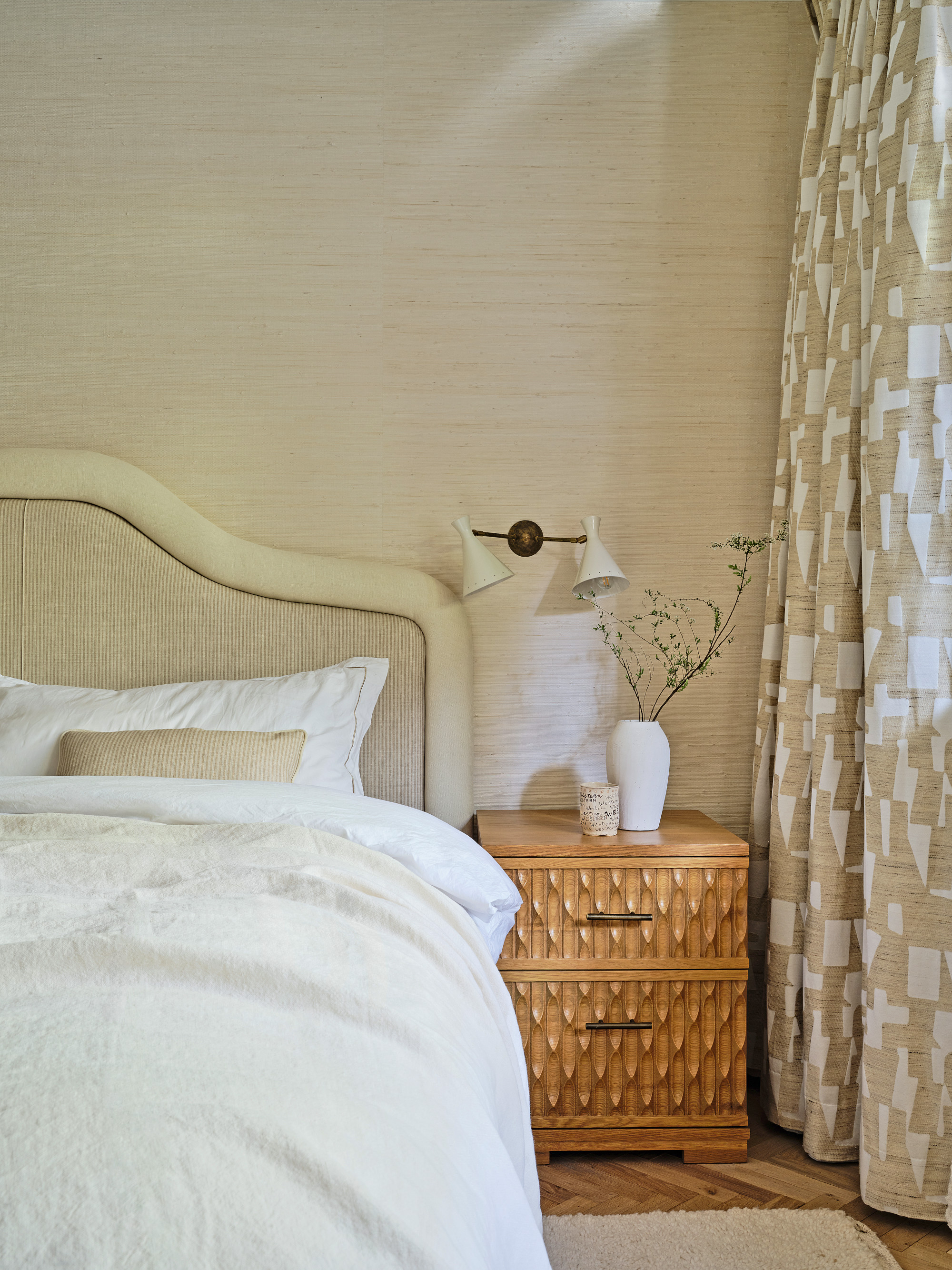 Beige bedroom with grasscloth wallpaper, cream upholstered headboard, abstract beige and white curtains, carved textured beside table and white bed linen