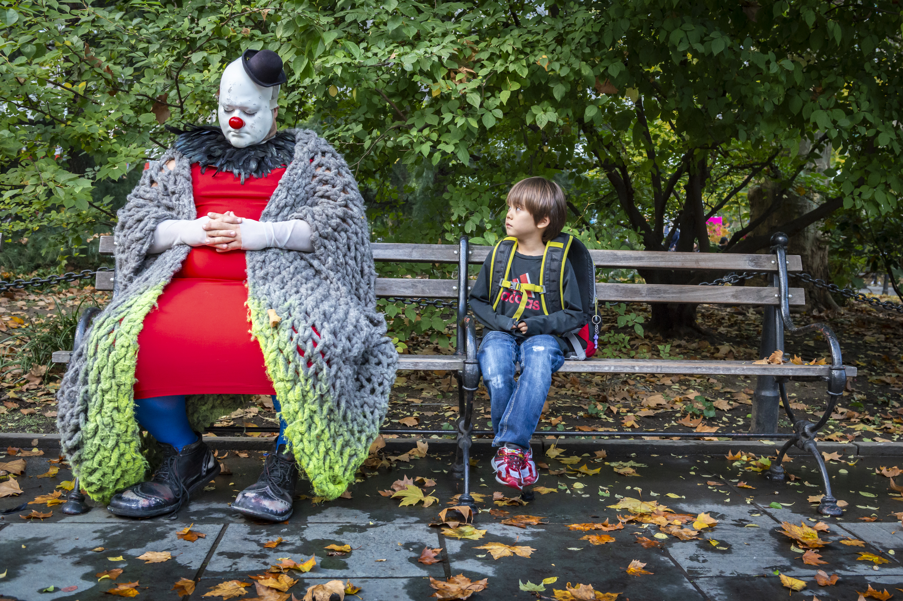 A child sits on a park bench covered in fallen leaves, looking curiously at a clown in a red dress and heavy knit shawl who is resting with closed eyes.