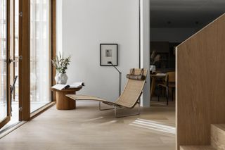 Modern residential interior featuring a curved lounger and side table in light natural colours. The walls are pale and complimented by the wooden staircase and wooden framed large windows.