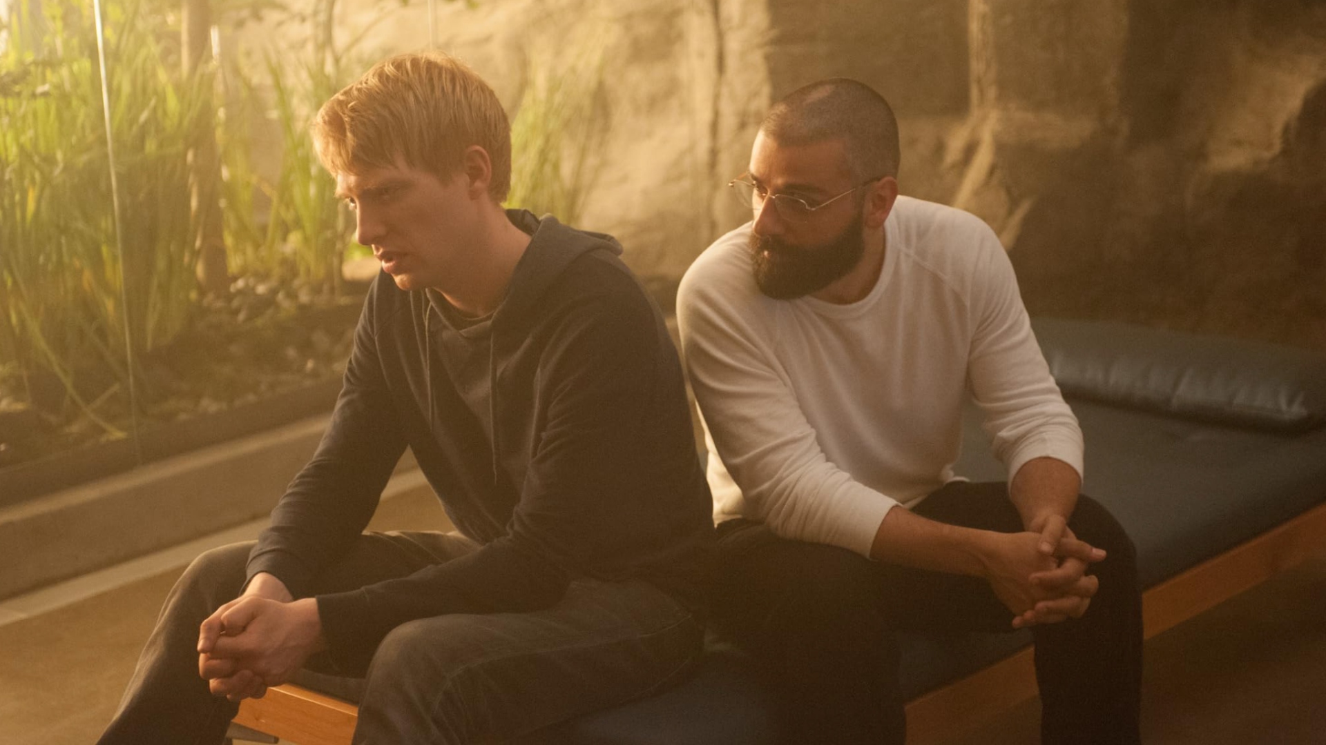 Two men sat together on a bed. The man on the left is blonde and clean shaven, and he looks upset. The man on the right has short hair and a full beard. Greenery can be seen outside the full-length window behind them.