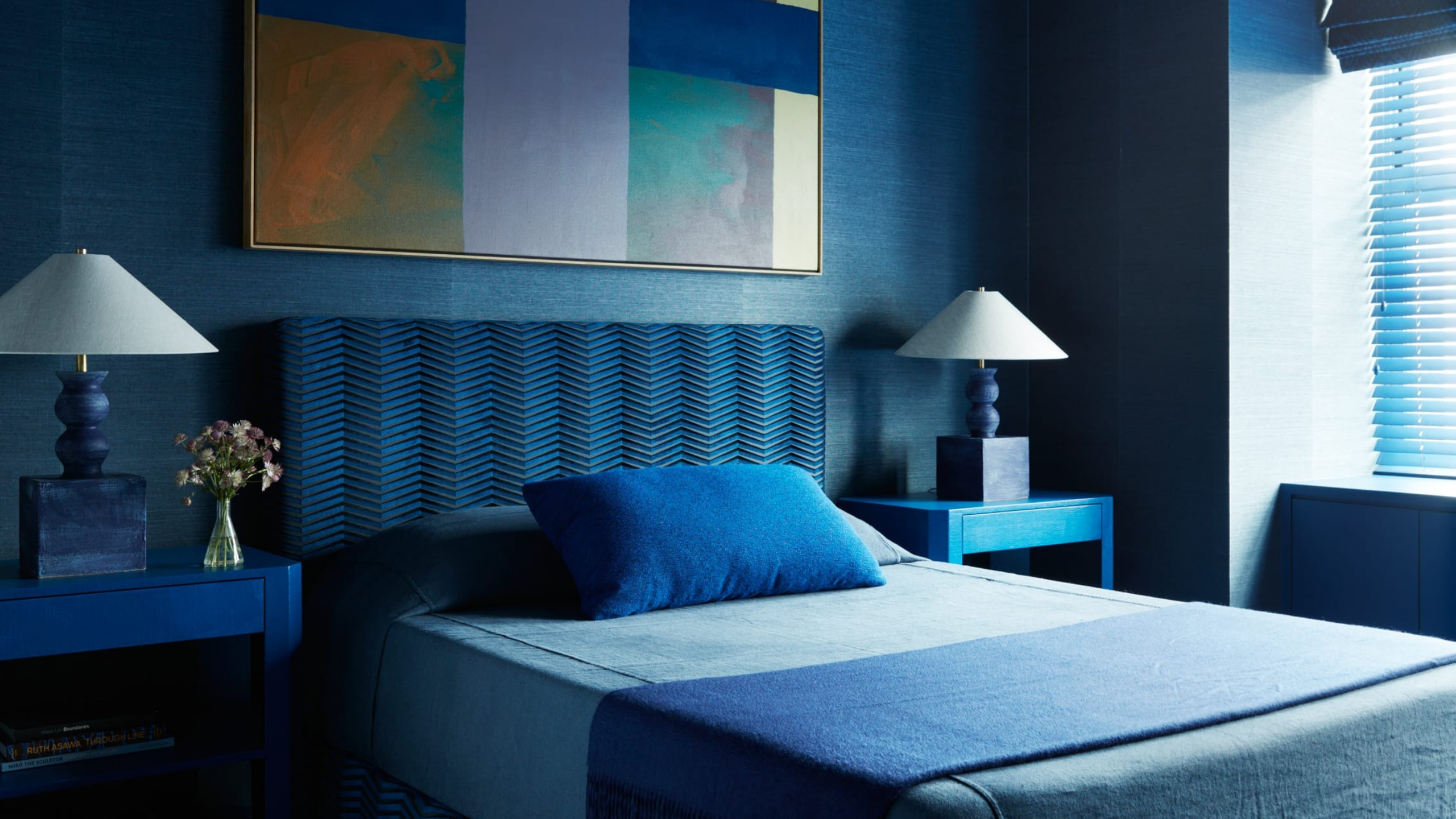 Blue bedroom with textured blue walls, matching upholstered headboard, layered blue bedding, sculptural bedside lamps, and abstract artwork.