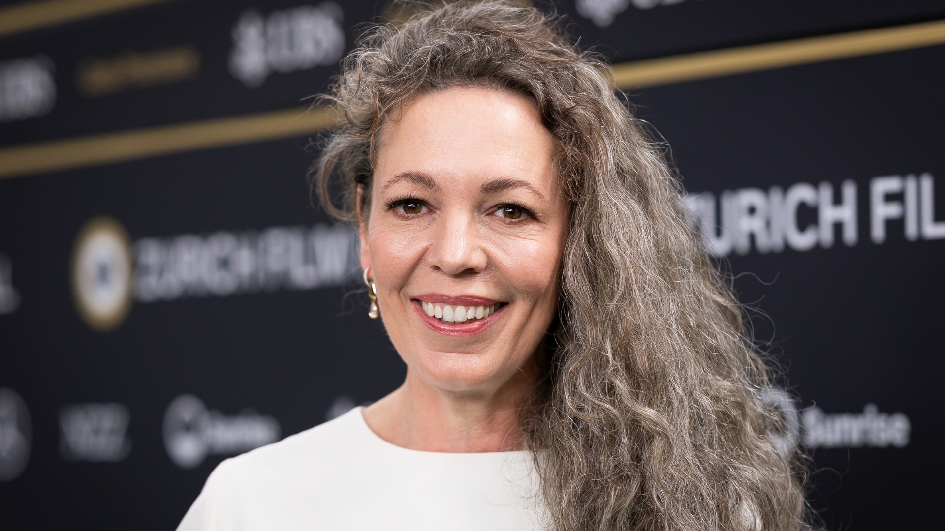 Image of Olivia Colman smiling with grey curly long hair over one shoulder