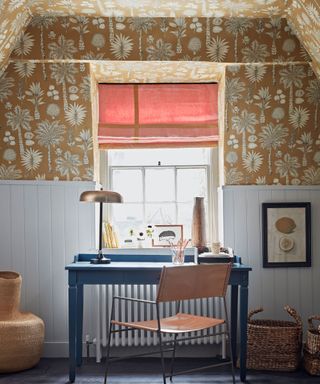 Contemporary study space with white and gold floral wallpaper on upper walls and ceiling, light blue paneling on low half of walls, blue wooden desk, brown leather and black metal frame chair, natural textured rug, woven baskets on floor
