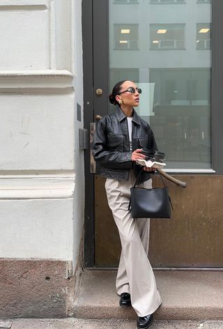 a photo showing what shoes to wear with wide-leg pants with a woman standing in a doorway wearing a leather jacket over a white t-shirt with tan wide leg trousers and black leather loafers