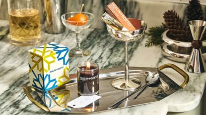 A dark rum scented candle on a chrome tray by a coupe with a vial of fragrance oil on a marble counter by a chrome jigger and a bowl of pinecones