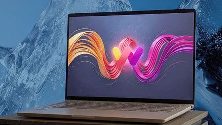 An ASUS Zenbook A14 on a wooden surface displays artwork featuring a stylized Windows Central logo surrounded by flowing ribbons. Behind the laptop is a textured blue background resembling ice.