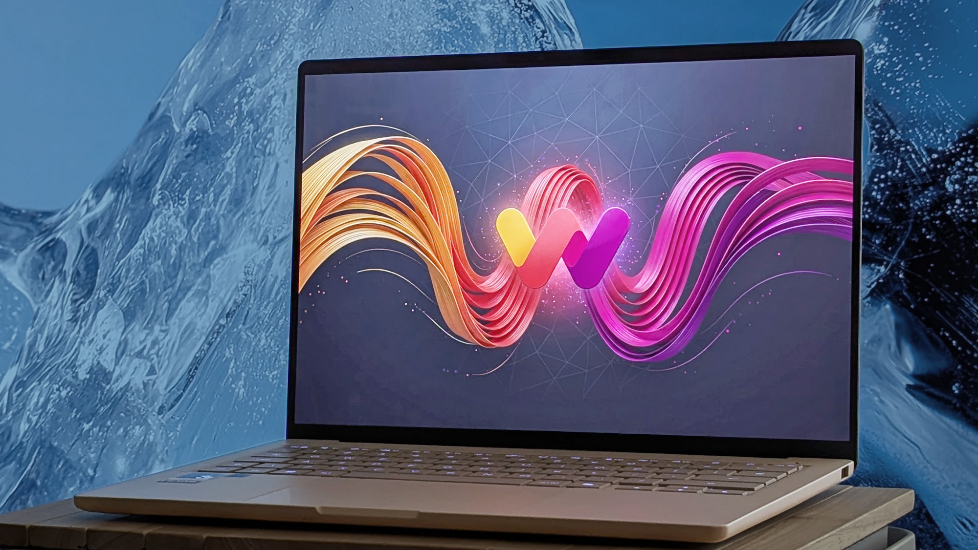 An ASUS Zenbook A14 on a wooden surface displays artwork featuring a stylized Windows Central logo surrounded by flowing ribbons. Behind the laptop is a textured blue background resembling ice.