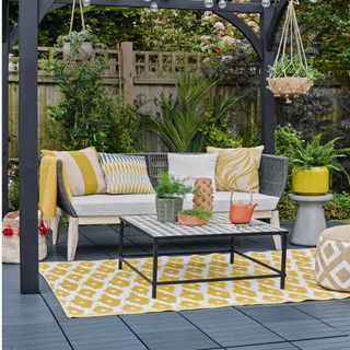 Black pergola with strings of light bulbs, sofa with cushions, rug and table with plants in pots