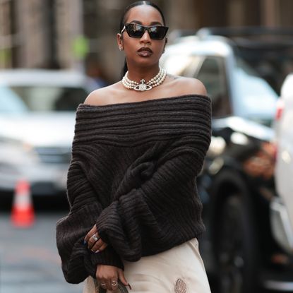 Fashion week guest is seen wearing an off-the-shoulder dark brown chunky knit sweater over a light cream-colored silky slip dress with a high slit and lace trim. She carries a brown leather handbag with a buckle in her left hand, while her right hand features rings and blue-toned nail polish. Accessories include a pearl Vivienne Westwood necklace and large black sunglasses, with her hair slicked back on Lapointe Show September 13, 2025 in New York City.